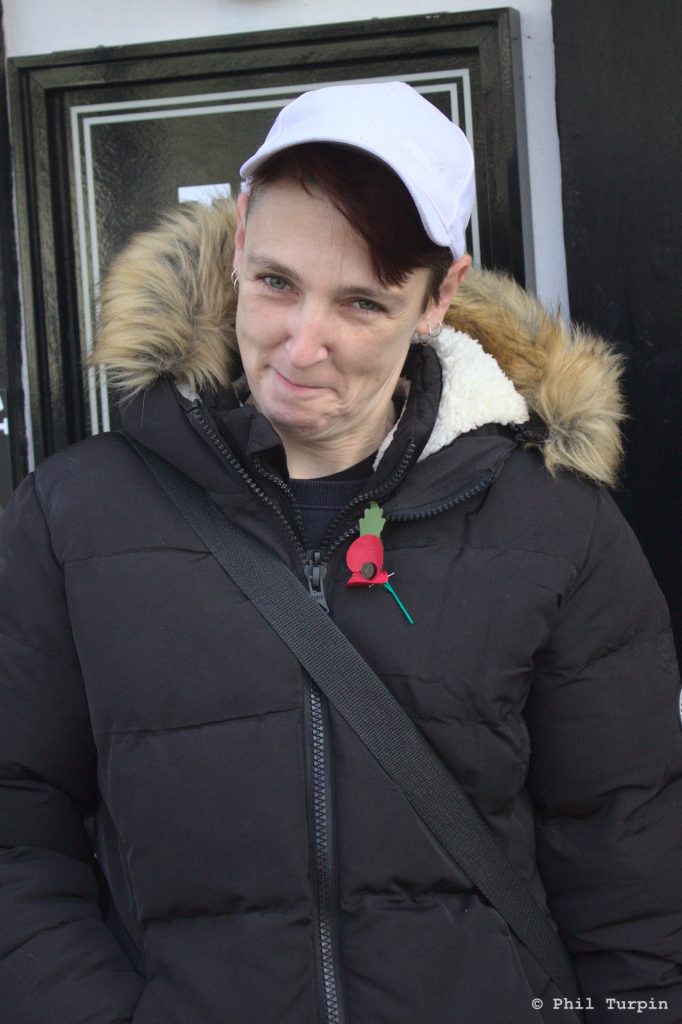 Street portrait of a person, wearing a white baseball cap, in a black parker jacket with fur hood, standing outside a pub in Ashby De La Zouch