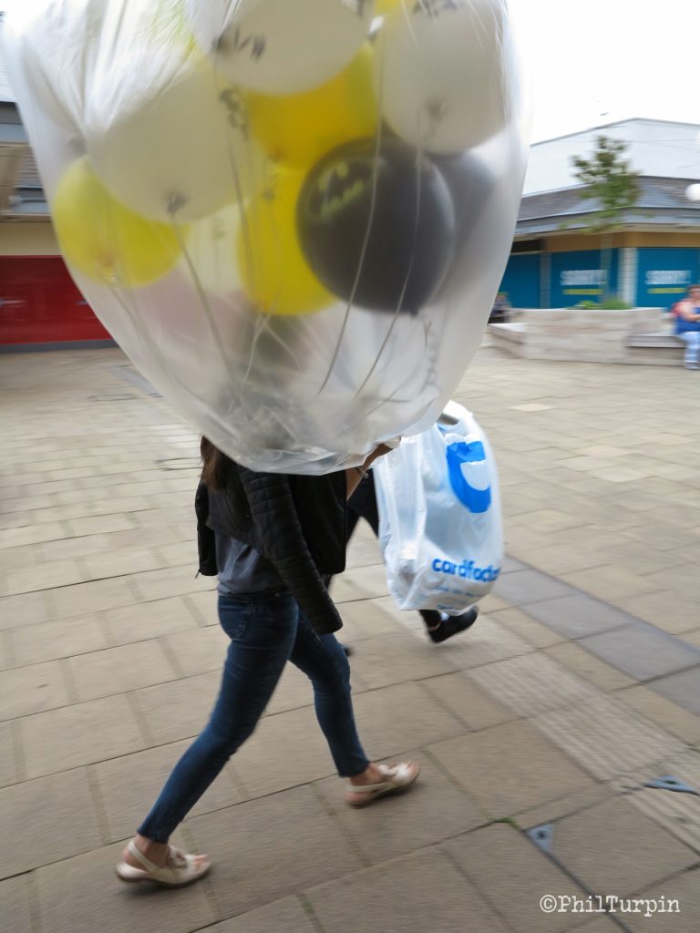 A person is close in frame, walking left to right, and holding a load of white, yellow, and black helium filled balloons in her hand. There is a big see through plastic bag containing all the balloons. She's also carrying a small white plastic bag with blue writing. There is open space in the background, of a shopping centre, that is slightly motion blurred.