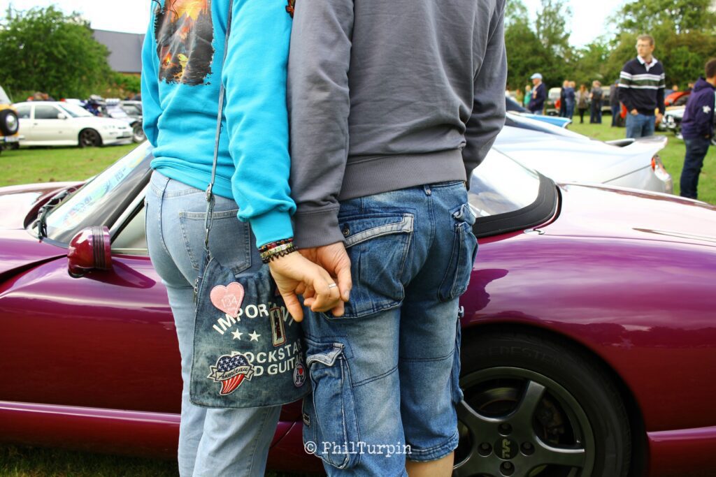A couple with their back to the camera, hold hands, while standing at the side of a purple TVR Chimeara. They're dressed in demin shorts and jeans, and one has a denim back with embroidered patches and badges. Their head's and feet are cropped out of frame.