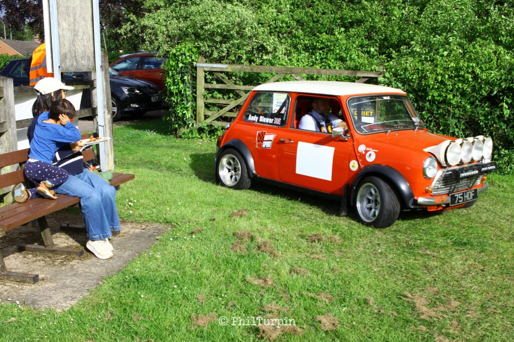 A classic red mini, with a white roof, for 4 spotlights on the bonnet, and rally decals, drives through a gate, in a field, while a mother and child, dressed in blue, sit on a bench to the left. The child has their hands over their ears because the car's got a loud exhaust.