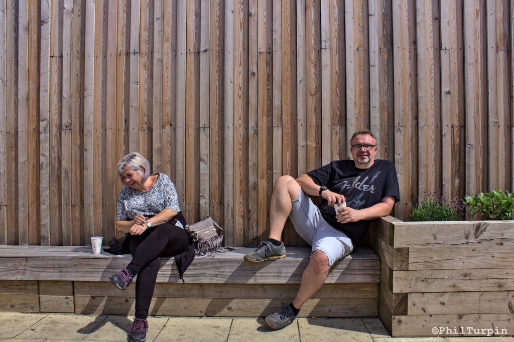 Two people sat on a bench with a planter to the right and wooden boards as the background. The person to the left has crossed legs and is looking away from the camera, the person to the right is looking straight at the camera, has one foot on the bench with his knee bent and is resting his elbow on the planter.