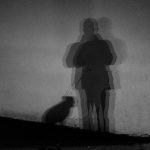 A black and white silhouette, against a light coloured wall, of a person standing with a dog next to them. There are three light sources from behind which gives the impression of two further outlines.